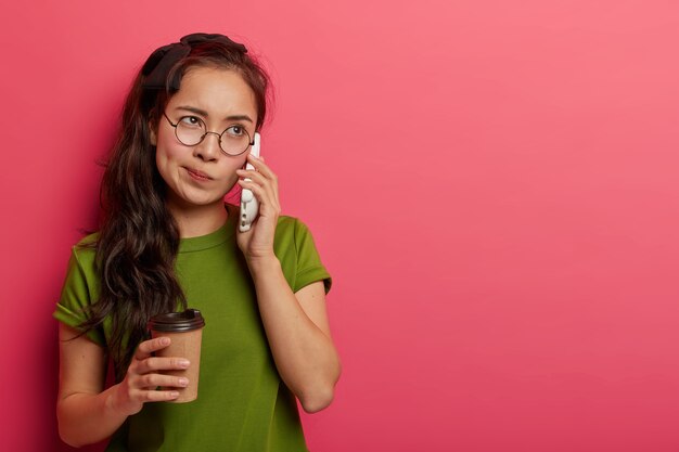 Millennial brunette female makes phone call, tired of waiting someone answers, keeps cellular near ear, drinks takeaway coffee, dressed in casual green t shirt, poses indoor.