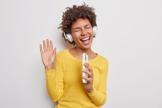 Foto gratuita la ragazza millenaria con i capelli ricci ascolta la musica tramite le cuffie stereo tiene lo smartphone come se il microfono godesse della canzone preferita utilizza l'applicazione musicale indossa un maglione giallo isolato su sfondo bianco