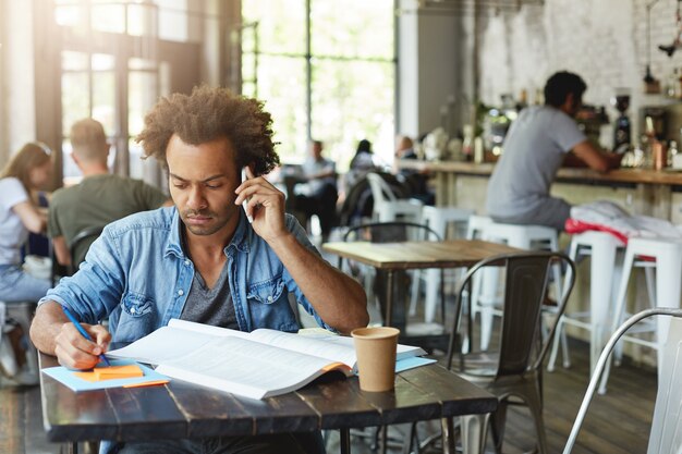Внимательный темнокожий студент-мужчина в повседневной одежде готовится к экзаменам, сидя за столиком в кафе, читая информацию в учебнике и разговаривая по телефону