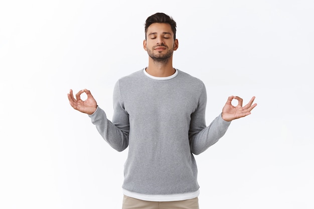 Бесплатное фото Внимательный терпеливый и расслабленный молодой счастливый человек, практикует дыхательные упражнения, держится за руки боком и с облегчением улыбается, снимает стресс во время работы, отдыхает, чтобы медитировать, белая стена