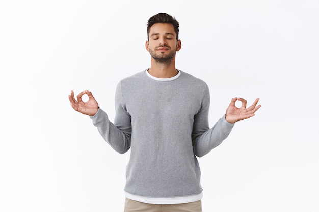 Free photo mindful patient and relaxed young happy man, practice breathing exercises, hold hands sideways and smiling relieved, release stress during work time take break to meditate, white wall