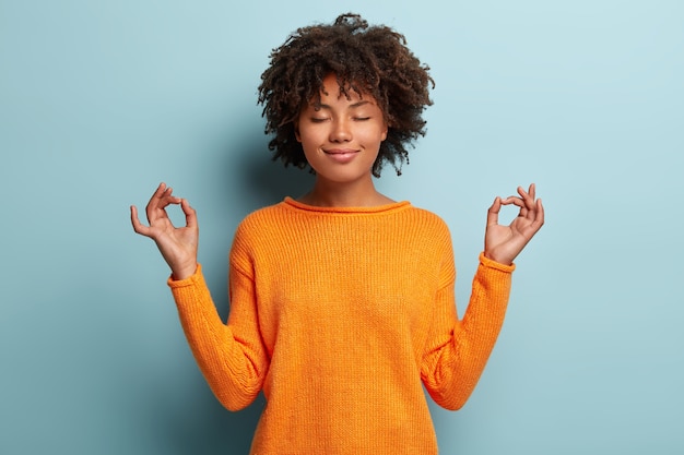 Mindful peaceful Afro American woman meditates indoor, keeps hands in mudra gesture, has eyes closed