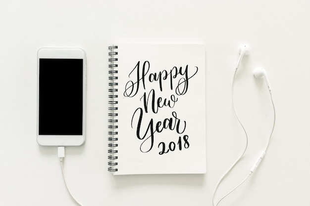 Minimal work space - Creative flat lay photo of workspace desk with "2018 Happy New Year" on sketchbook and mobile phone with blank screen and earphone on white background. Top view, New year concept.