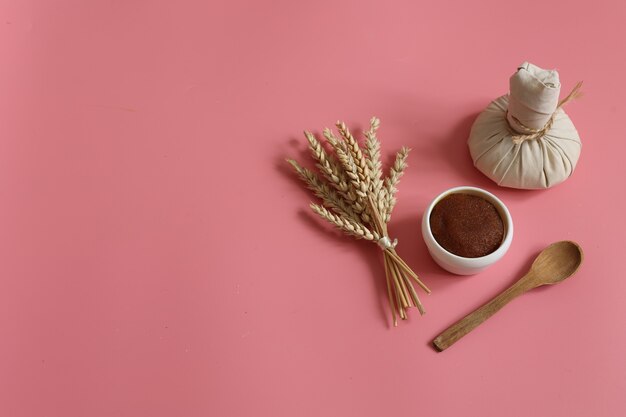 Minimalistic spa composition with natural scrub, herbal massage bag, wooden spoon and wheat on a pink background, copy space.