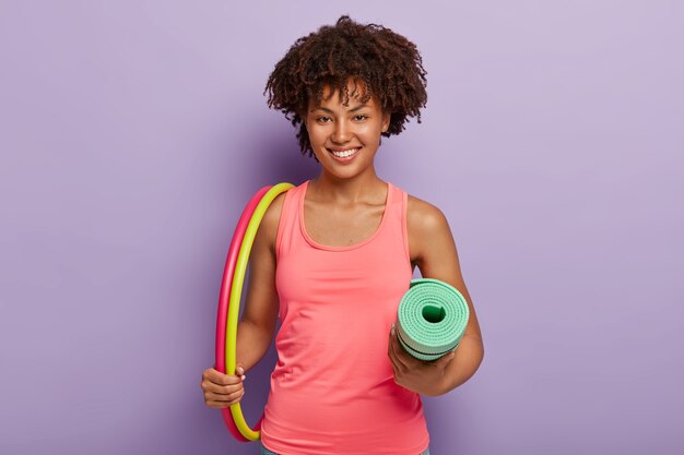 Веселая афроамериканка несет два обруча, свернул каремат, делает упражнения для похудания