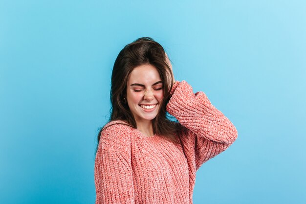 장난 꾸러기 소녀는 파란색 벽에 귀여운 표정을 만듭니다. 따뜻한 스웨터에 검은 머리 여자가 웃음.