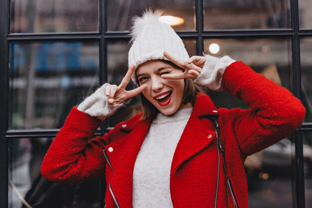 Mischievous short-haired girl in warm hat and red coat winks. Shot of woman with red lipstick showing peace signs against window.