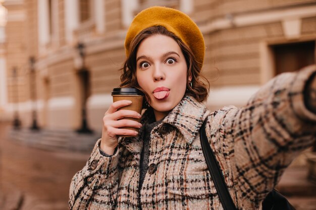 Mischievous woman in autumn outfit makes selfie against wall of city