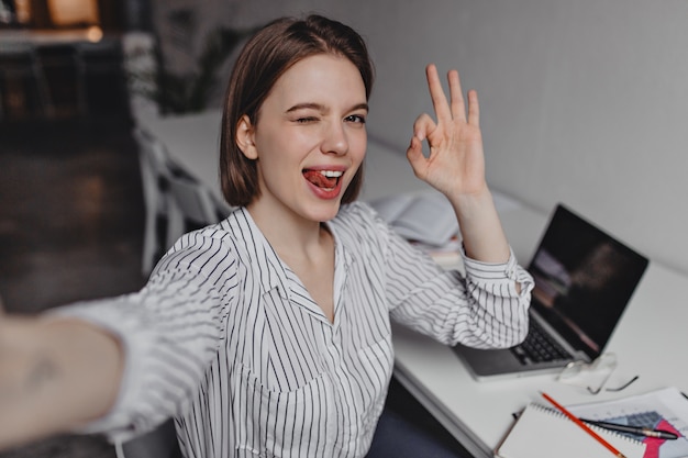 Бесплатное фото Озорная женщина делает селфи в офисе. офисный работник в полосатой рубашке показывает знак ок на фоне ноутбука.