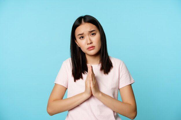 Miserable asian girl asking for help, pleading, begging for smth, standing sad and gloomy against blue background.