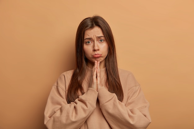 Miserable gloomy woman pleads for mercy, keeps palms together, makes apologize, purses lower lip, hopes or wishes something, stands with prayer gesture