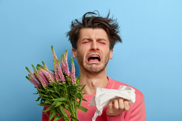 Miserable man in depression suffers from allergic malaise and rhinitis, seasonal disease, tired of sneezing, has red nose and eyes, allergy to flowering, holds handkerchief, feels irritation