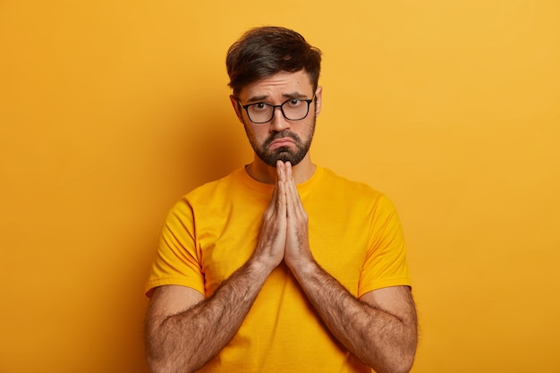 Miserable sad pleading man says sincere please, makes apology, keeps palms pressed together, begs with upset expression, needs help in trouble, prays with hope has faith in better wears yellow t shirt