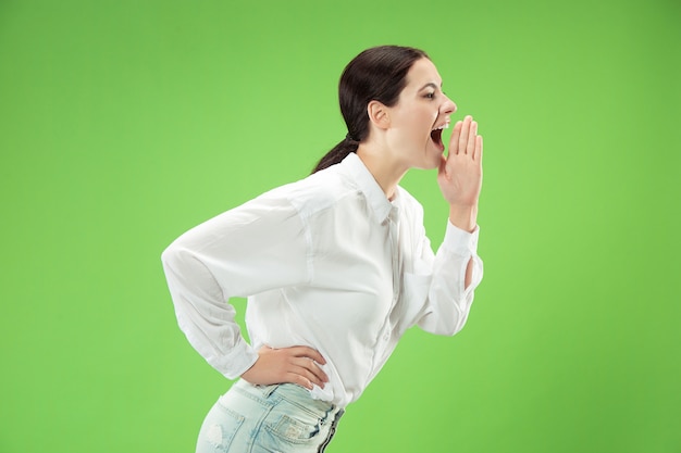 Do not miss. Young casual woman shouting. Shout. Crying emotional woman screaming on green studio background. Female half-length portrait. Human emotions, facial expression concept. Trendy colors