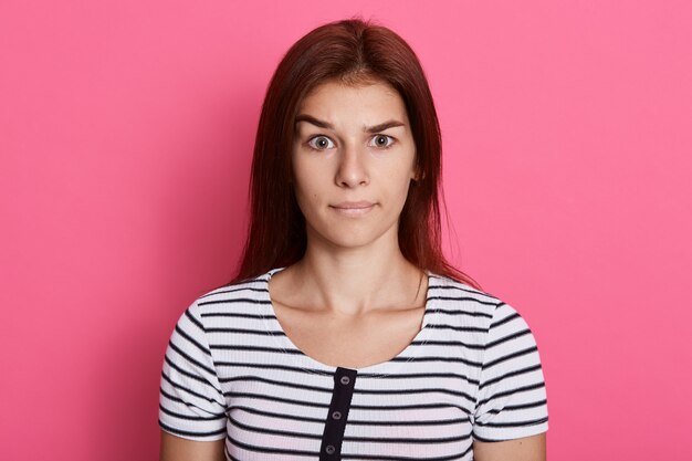 Misunderstanding lady , wearing casual striped t shirt, posing isolated over pink wall,  with serious look.