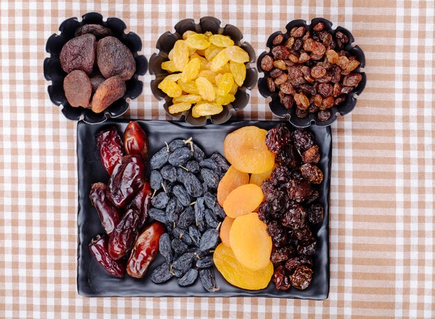 Mix of dried fruits dates raisins apricots and cherries on a black tray and in mini tart tins on plaid tablecloth top view