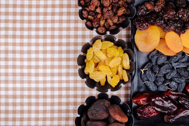 Mix of dried fruits dates raisins apricots and cherries on a black tray and in mini tart tins on plaid tablecloth with copy space top view