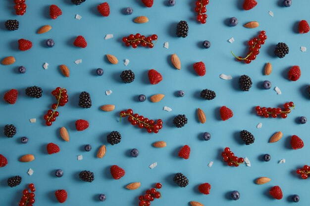 Mixed assorted fresh berries including raspberry, blueberry, blackberry, redcurrant on blue background. Almond nuts and coconut flakes. Healthy ingredients for making tasty dessert. Big variety