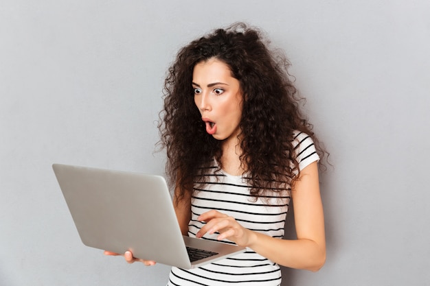 Mixed emotions of beautiful woman being surprised or outraged while chatting in social network using personal computer over grey wall
