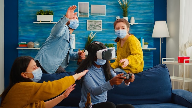 Смешанная группа людей ведет чернокожую женщину с гарнитурой VR, играющую в виртуальные видеоигры в гостиной, сохраняя социальное дистанцирование от covid19. Разнообразные друзья веселятся на новой нормальной вечеринке.