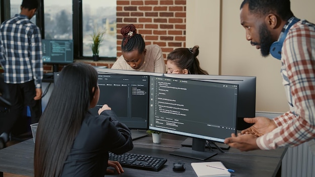 시작 사무실에서 실행 중인 코드를 보여주는 여러 화면에서 그룹 프로젝트 작업을 하는 혼합 프로그래머 팀. 소프트웨어 컴파일 코드가 있는 컴퓨터 화면에 연필을 가리키는 코더.