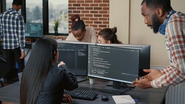 시작 사무실에서 실행 중인 코드를 보여주는 여러 화면에서 그룹 프로젝트 작업을 하는 혼합 프로그래머 팀. 소프트웨어 컴파일 코드가 있는 컴퓨터 화면에 연필을 가리키는 코더.