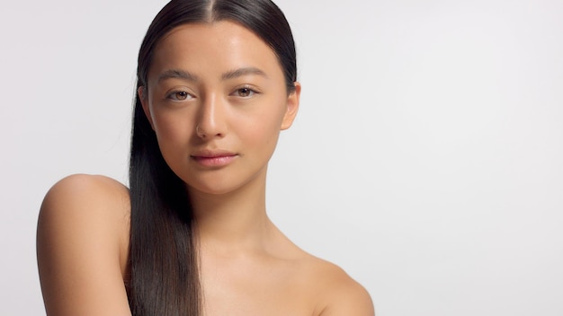 スタジオビューティーシュートの混血アジア人モデルモデルがカメラにポーズをとるストレートヘアを右側にコーミング理想的なskniとメイクアップなし頭と肩のクロップカメラを見る