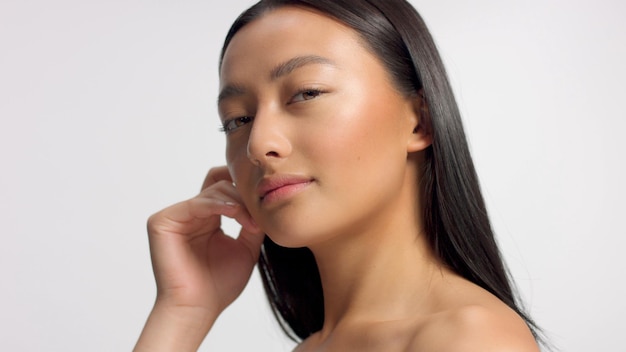 Mixed race asian model in studio beauty shoot Model poses to a camera straight hair Ideal skni and no makeup makeup Head and shoulders crop touches her skin with hand