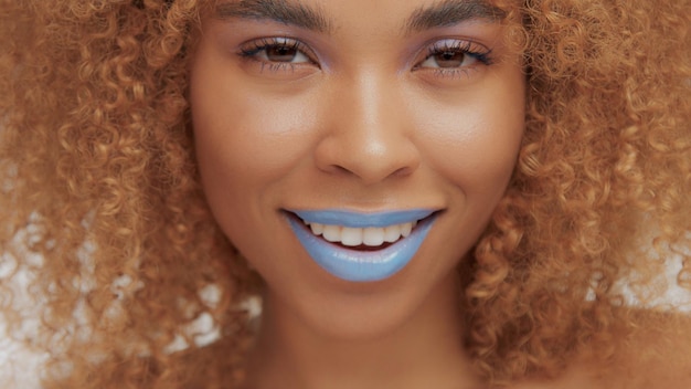 Mixed race black blonde model with curly hair closeup of mouth painted with bright blue lipstick smiling and watching to the camera