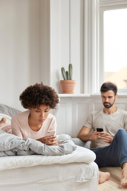 Mixed race boyfriend and girlfriend read recieved notification, send sms while rest in bedroom, ignore live communication, have serious expressions, focused in cellular. Addiction and technology