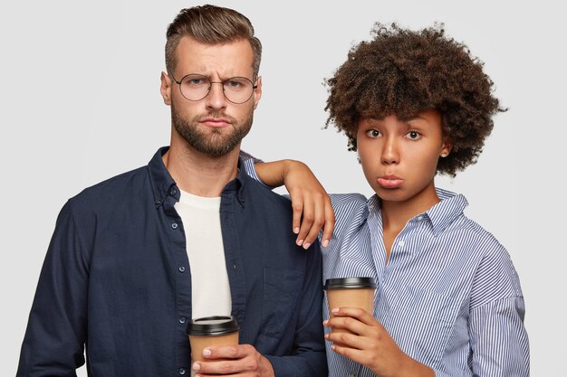 Mixed race female and male student have dissatisfied expressions, drink coffee after lectures, discontent with exam results. African American woman leans at shoulder of companion, stand together