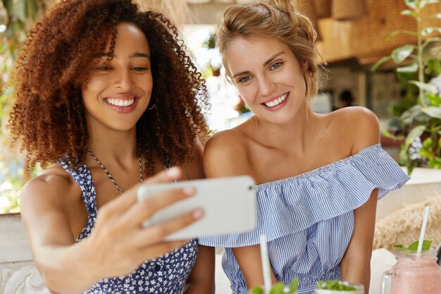 Mixed race friendly females have positive expressions, pose for selfie in modern smart phone, have satisfied looks, recreat in coffee shop together. African American woman takes photo on cellular