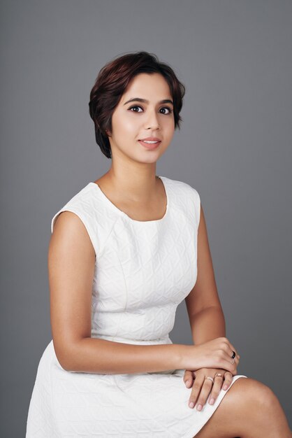 Mixed race lady in dress sitting in studio with crossed legs, posing and smiling