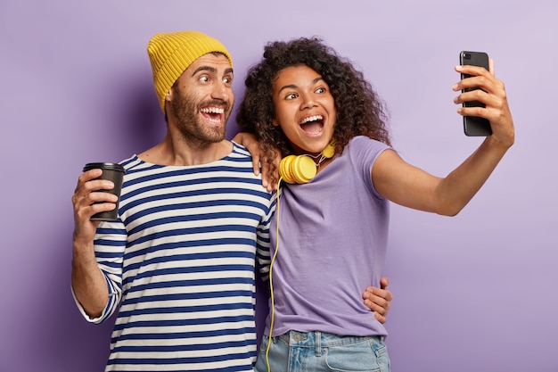 Mixed race lovely couple embrace and stand closely, pose for making selfie portrait, have glad expressions, drink coffee