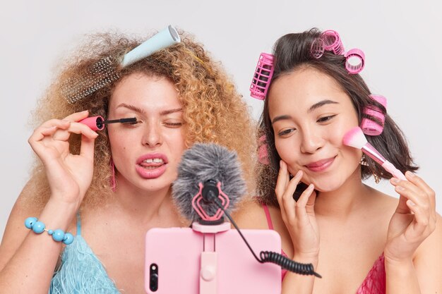 Mixed race professional women vloggers do make up beauty products review record video blog via smartphone give useful tips to subscribers film process of applying mascara and face powder do hairstyle