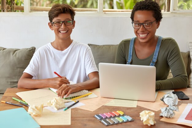Mixed race students of high school learn together at coworking space, watch training webinar on laptop computer, write records in spiral notebook, find creative solution, have toothy smiles.