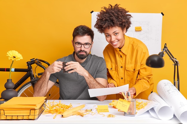 Free photo mixed race woman and man collaborate together discuss future project and try to find best variant for sketches pose at messy desktop
