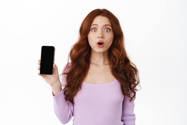 Mobile advertisement. Excited redhead woman gasp wow, shows smartphone empty screen, stare impressed at camera, special deall online shopping, standing against white background