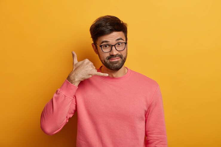 Free Photo mobile communication concept. unshaven man makes call me gesture, talks on imaginary cellular, has friendly positive expression, wears transparent glasses and pink jumper, isolated on yellow wall