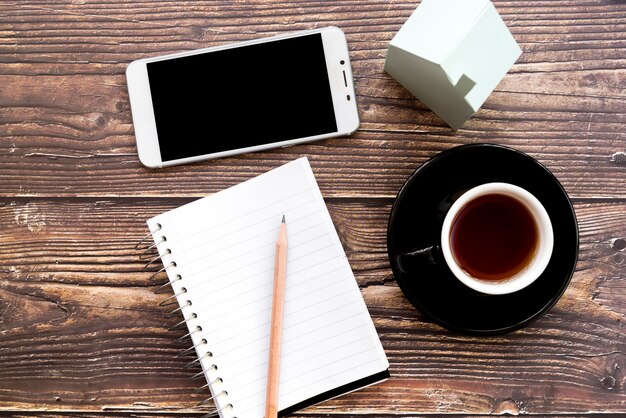 携帯電話;空白のスパイラルノート。鉛筆;木製の机の上のコーヒーカップと家のモデル