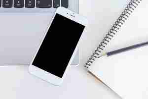 Free photo mobile phone over the laptop with pencil and notebook over the white table