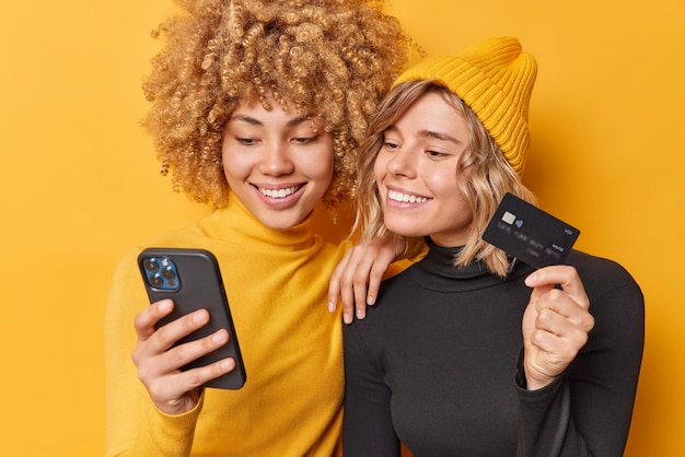 Mobile shopping and banking concept positive glad young women use credit card and smartphone application make online purchases dressed in casual turtlenecks isolated over yellow background Premium Photo