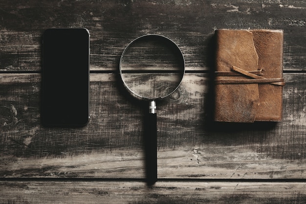 Mobile smartphone, magnifier and notebook with leather cover isolated on black farm wooden table Mysterious detective game concept.