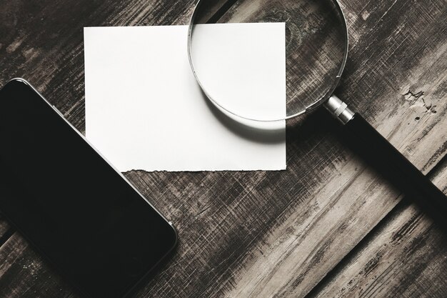 Mobile smartphone, magnifier and sheet of white paper isolated on black farm wooden table Mysterious detective game concept. Closeup side view