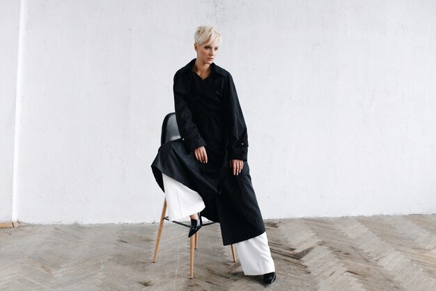 Model in black coat sits on a bar chair before a white wall thoughtful