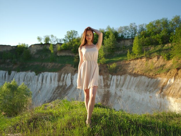 Model posing on a sunny day with a great sunny landscape around her. Young woman standing by a cliff with a nice view behind he back. Attractive girl with a white dress posing outdoors.