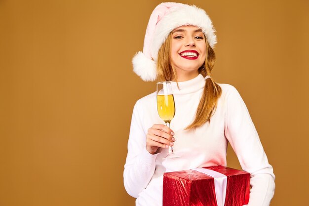 Модель в шляпе санта-клауса и большой подарочной коробке пьет шампанское на золотой стене