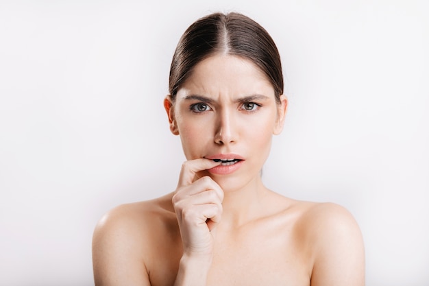 Model without makeup posing on white wall. Portrait of displeased woman biting from doubt finger.