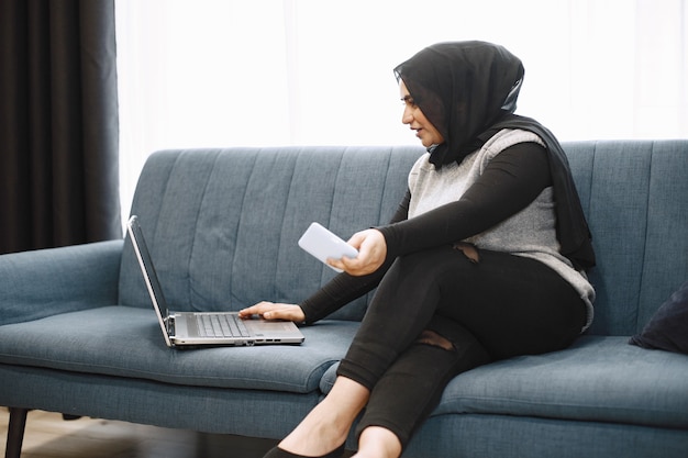Современная арабская девушка в хиджабе, используя ноутбук дома, работая удаленно, сидя на диване в гостиной
