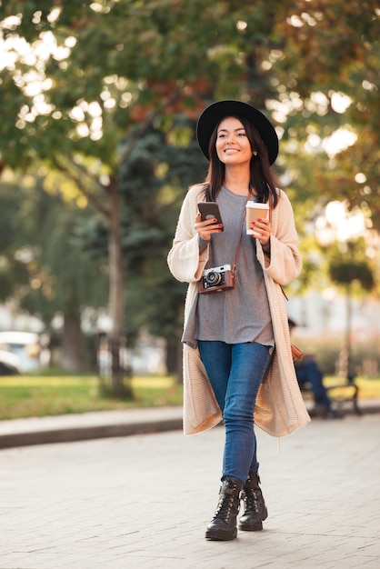 屋外の公園を歩きながら携帯電話とコーヒーカップを保持している現代のアジアの女性